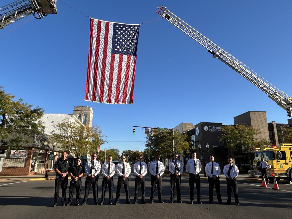 A group of firefighters in uniform stands on a city street. A large American flag hangs above them.