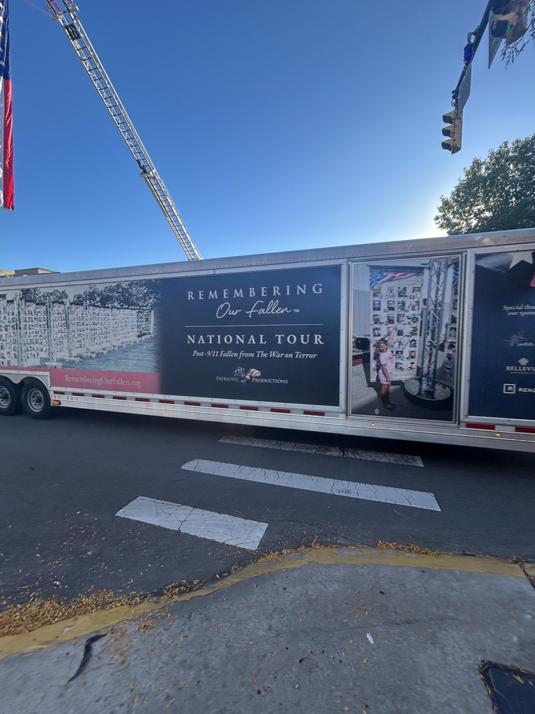 A large trailer with "Remembering Our Fallen" on the side, with an American flag behind it, is parked on a road.
