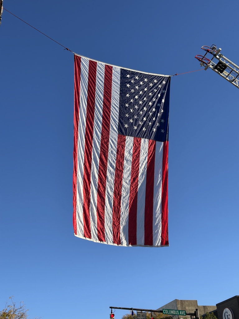 American flag suspended between two buildings, attached to wires. A firetruck ladder is partially visible above the flag.