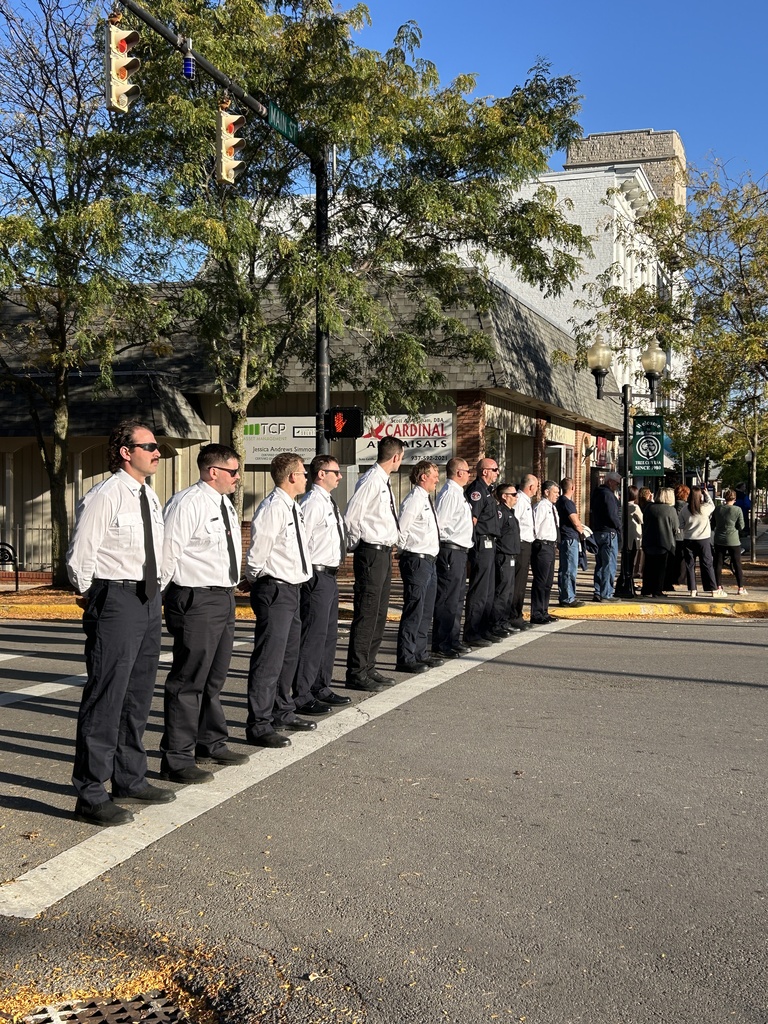 A group of men in uniform stands at a crosswalk, facing left, with buildings and trees in the background.