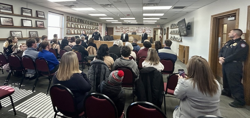 Audience seated in a meeting room faces a panel of speakers at the front. Walls display numerous framed photos. Police officers present. Engaged atmosphere.