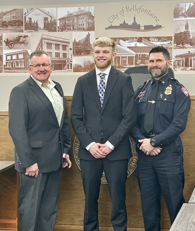 Three men stand in front of a "City of Bellefontaine" mural. The central man wears a suit, flanked by men in a blazer and police uniform, all smiling.