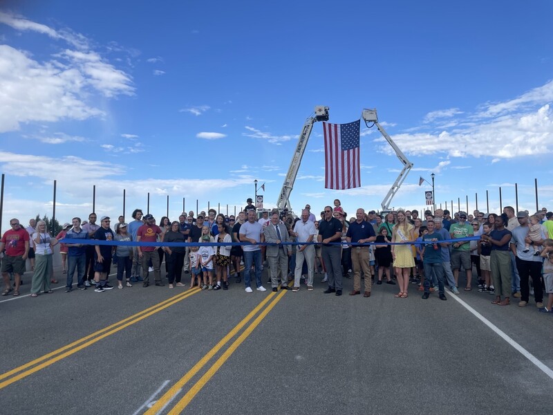 A crowd of people stand in front of an American flag suspended high between two ladder trucks. A blue ribbon is stretched out in front and one person hold large scissors and is prepared to cut the ribbon.