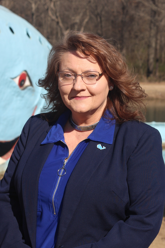 A women in a dark blue jacket and collared shirt, wearing glasses, standing in front of the Blue Whale attraction in Catoosa, OK.
