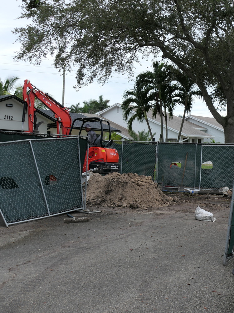 Backhoe removing dirt from work area surrounded by a green fence and home in the background.