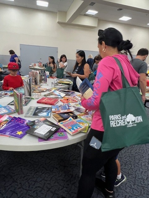 Image of books on a table and adults and children looking through books at our event called Book Swap