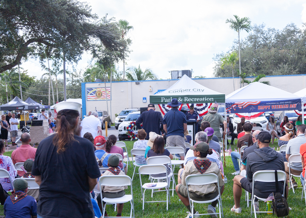 People standing and sitting in chairs with tens in the background, during Veteran Day ceremony.