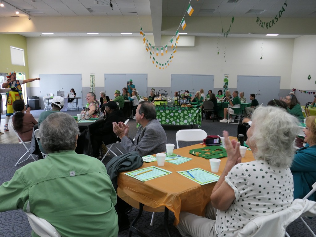 Room filled with people sitting at round tables with a man dressed as a clown pointing at someone inside the room.