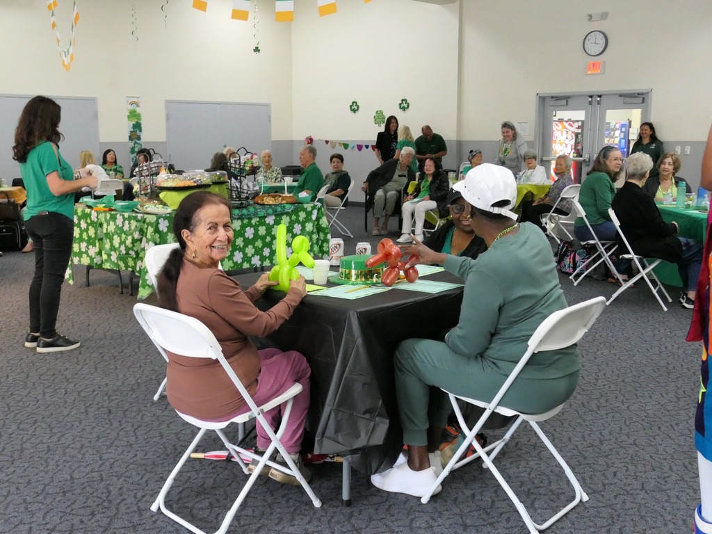 Woman sitting a table smiling as two other women hold balloons.