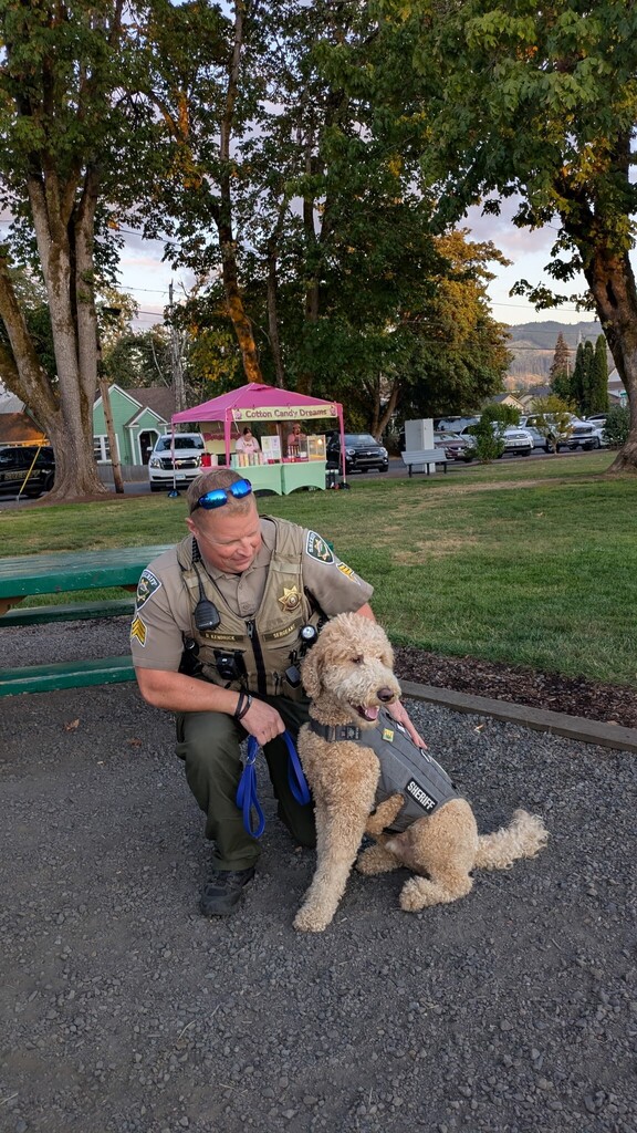 Image of Lane County Sheriff's Office Deputy Kendrick kneeling on the ground next to Corporal Carlton, the labradoodle that serves as the organization's Emotional Support Animal.