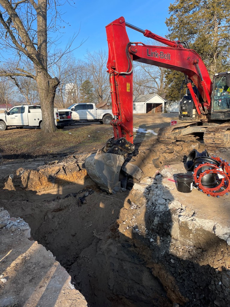 A red excavator digs a deep trench in a roadway during a water utility construction project. Broken concrete and soil surround the trench, with construction vehicles parked nearby and residential homes visible in the background on a clear day.