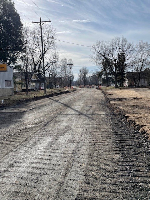 A residential street under construction with the pavement removed, exposing a rough, dirt and gravel surface. Deep tire tracks run along the roadway, and the edges are dug out on both sides. Orange construction barricades are visible in the distance blocking part of the road. Utility poles and overhead power lines line the street, with houses and leafless trees on either side under a partly cloudy sky.