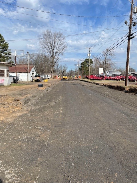 A partially paved road under construction stretches into the distance, with construction equipment ahead, utility poles and overhead wires lining both sides, a small building on the left, and rows of red equipment or machinery on the right under a partly cloudy sky.