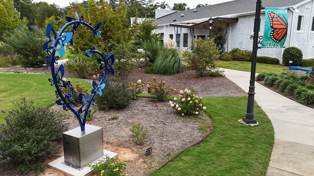 Left side shows a metal sculpture with a sliver base and a blue top with butterflies of many sizes along curved metal. it is surrounded by pollinator plants. to the right is a pole with a sign that says Center for the Arts with a butterfly on it