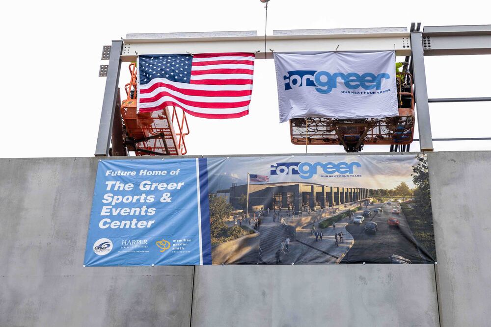 Image of concrete wall and beams with a banner on the wall that reads: "Future Home of The Greer Sports and Events Center" with a rendering to the left. Above it hanging on a white steel beam is an American flag and a white flag that reads "ForGreer"