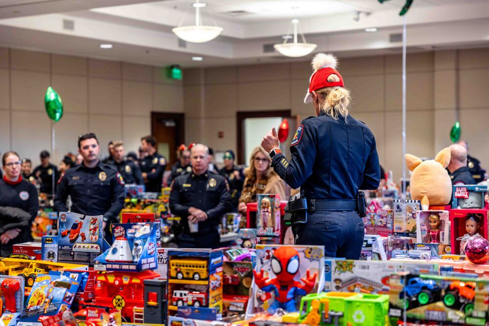 Hundreds of donated toys lined up on tables in the foreground and several Greer police officers listening to organizer speak in background