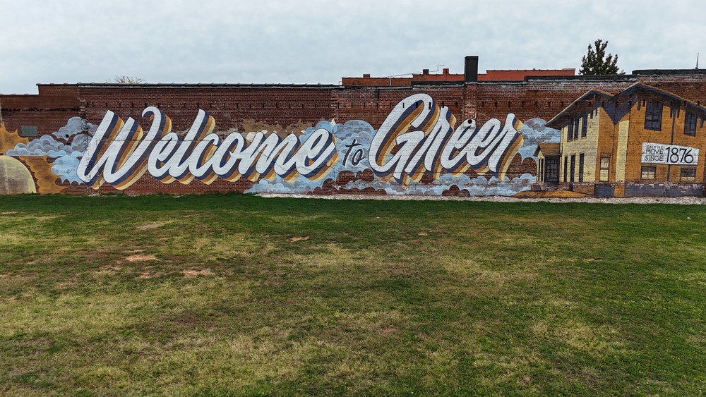 Side of a brick building where Welcome to Greer is painted in cursive along with the Historic Greer Depot. A grassy field is in front of the mural