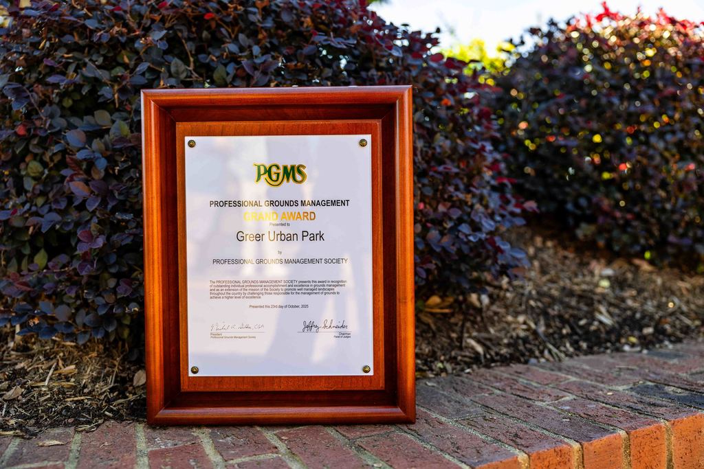 The PGMS Grand Award for Greer City Park sits on a brick half wall in Greer City Park with bushes behind it
