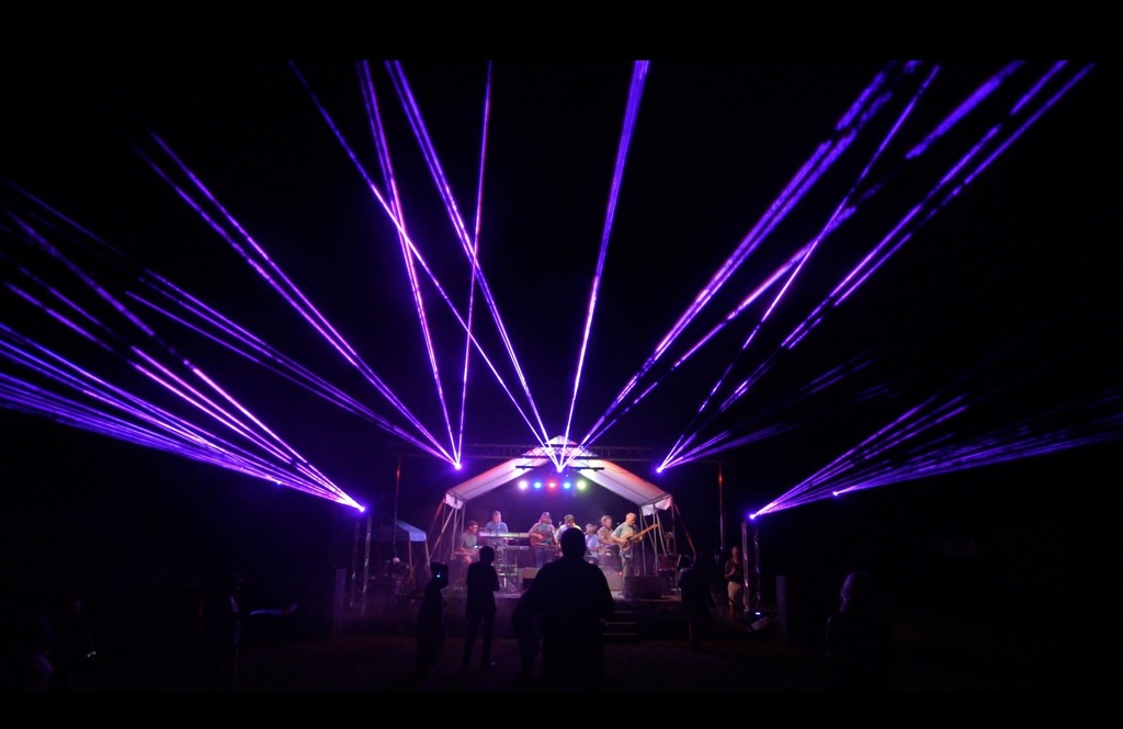 Multiple purple laser lights radiate out from a stage with onlookers in the crowd
