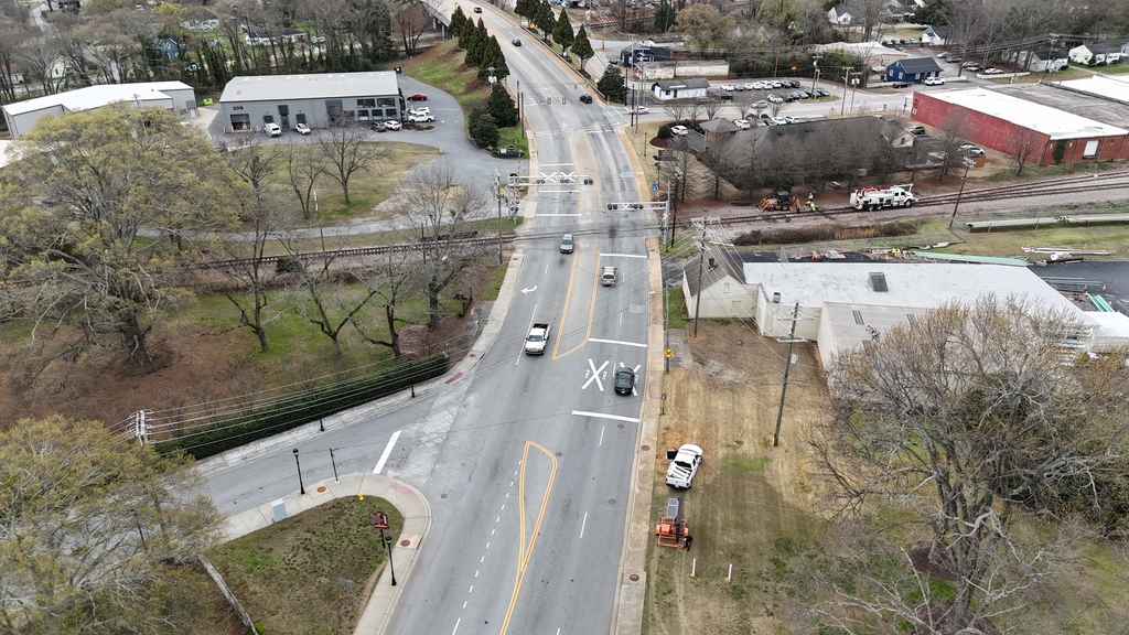 A drone shot of a four lane roadway with multiple cars driving down up and down it, with a train track running perpendicular to the road 