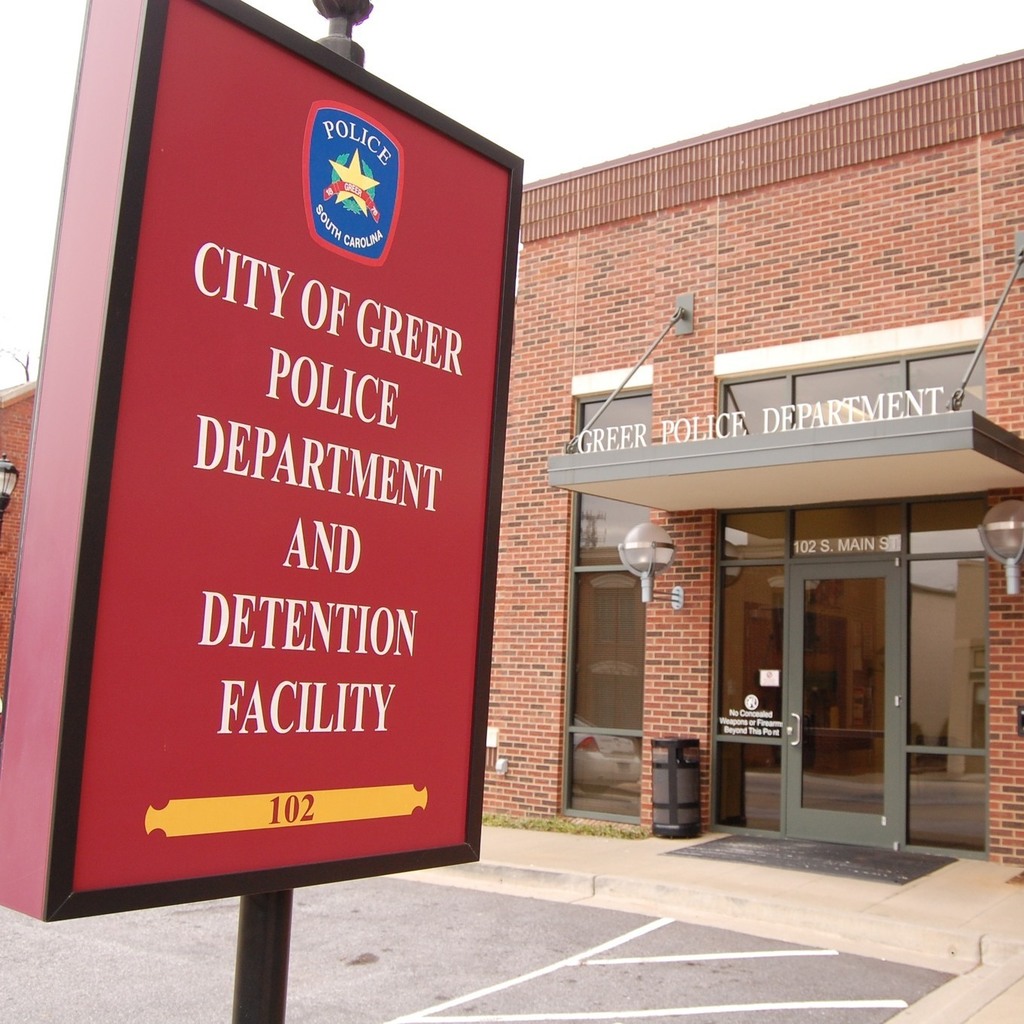 Close up of a red sign that reads City of Greer Police Department and Detention Facility with the Greer Police Department in the background