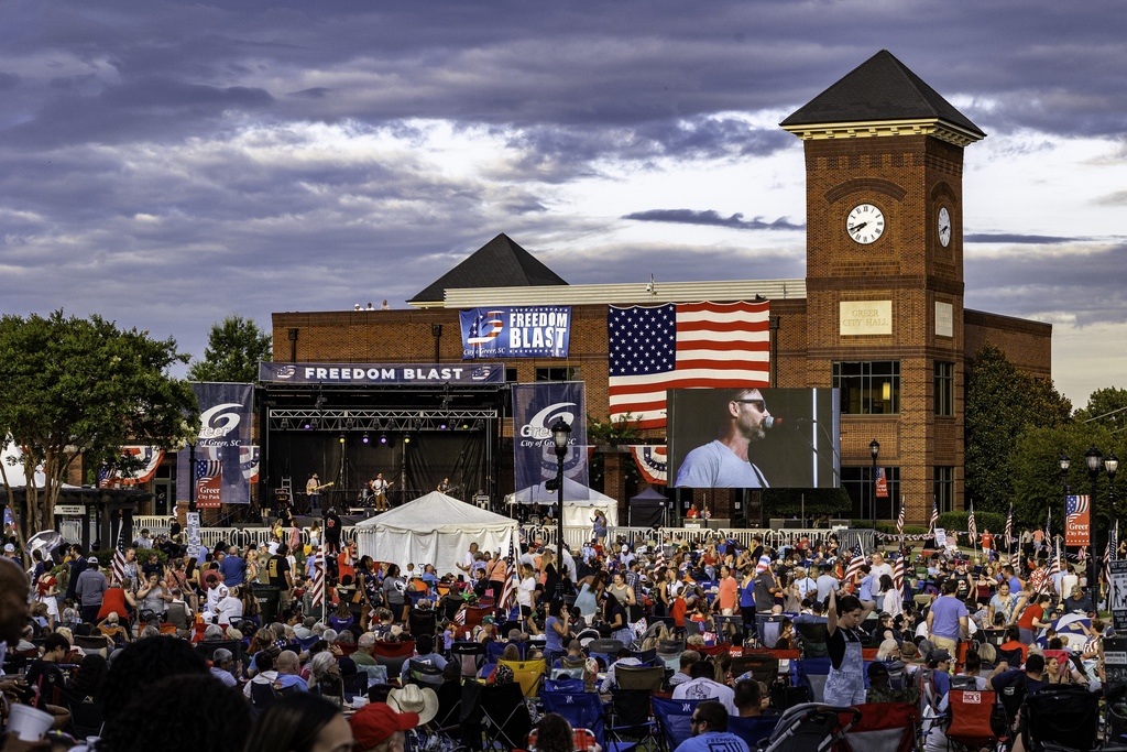 Picture shows thousands of people in Greer City Park watching a band play with an American flag draped over the back side of City Hall