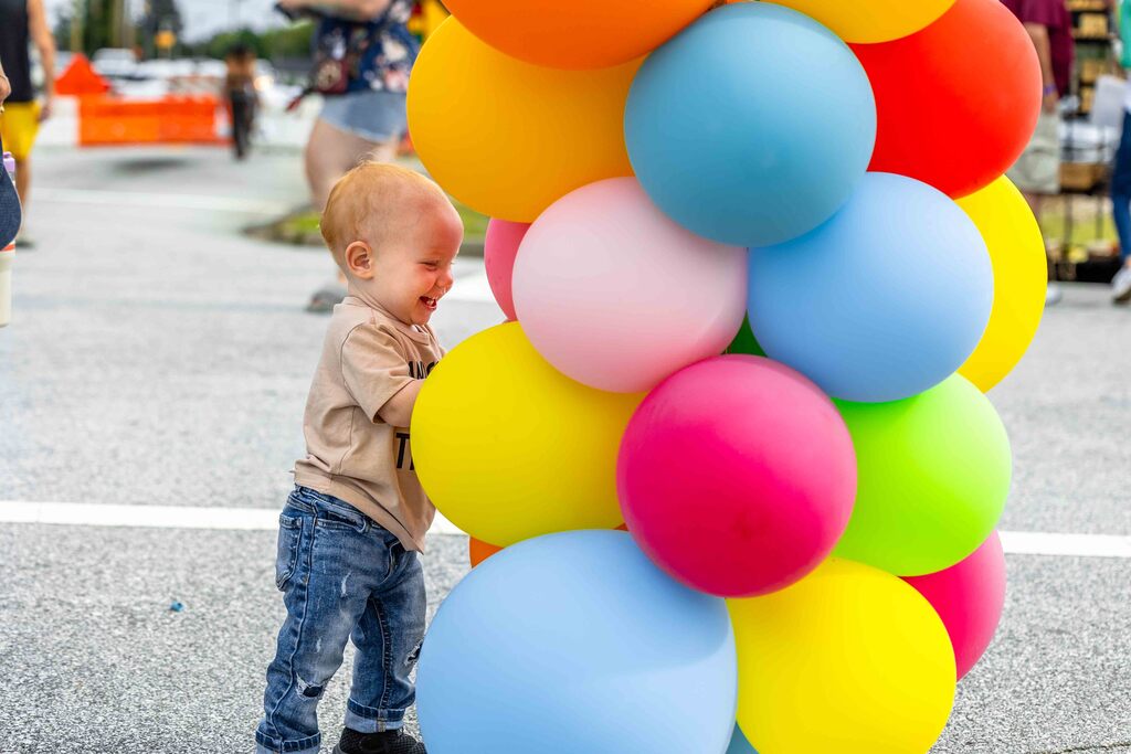 Toddler boy is smiling and touching an arch of colorful balloons on Depot Street in Greer