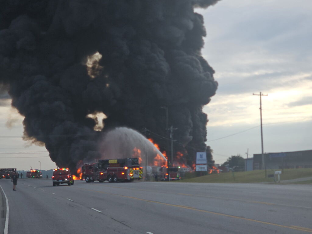 large plume of black smoke and multiple fire trucks putting water on the fire