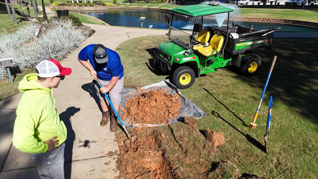 One city employee digging near a sidewalk in Greer City Park, another is standing by with hands on his hips, and a  green ATV and more shovels are on the right side