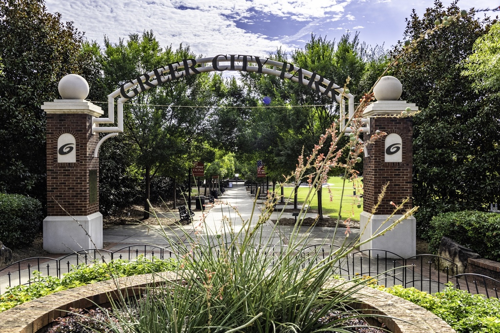 Circular brick flower bed in front with tall plants jutting out; background is the entrance to Greer City Park which has brick columns on either side and a metal archway in the center baring the park name in black lettering