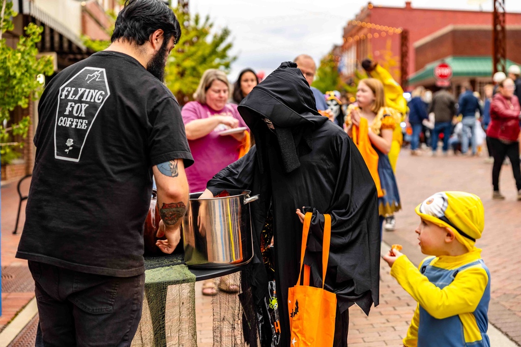 Man in black Flying Fox Coffee shirt handing out candy to a child in a black hooded costume and a toddler dressed like a yellow and blue minion