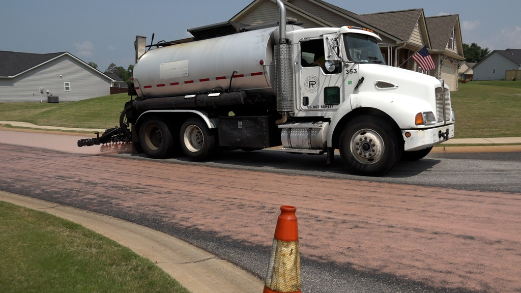 tanker truck spraying pink liquid on a road with house in background and traffic cone in front