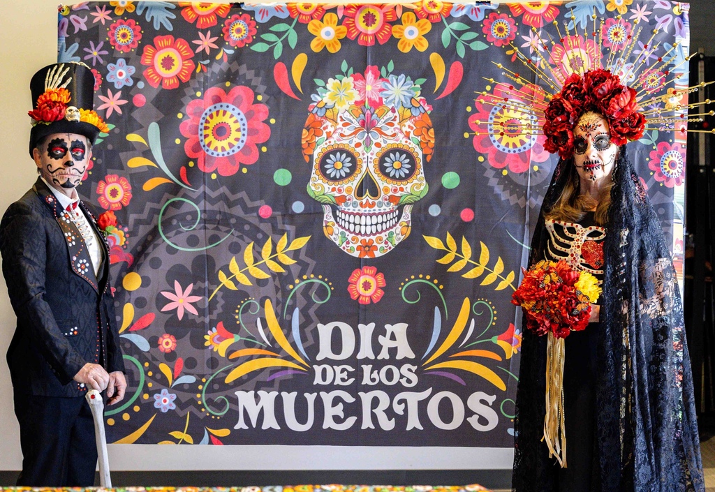 man and woman dressed up as a bride and groom in all black with faces painted standing on either side of a colorful banner that says Dia de los Muertos