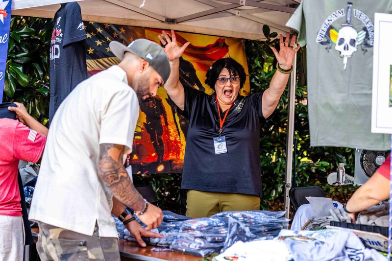 Person under tent smiles and waves at camera while people browse shirts