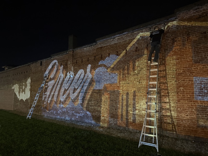 Picture shows a mural design that says Greer being projected on the side of The Great Bay Oyster House brick building and the artist tracing the projected image onto the wall.