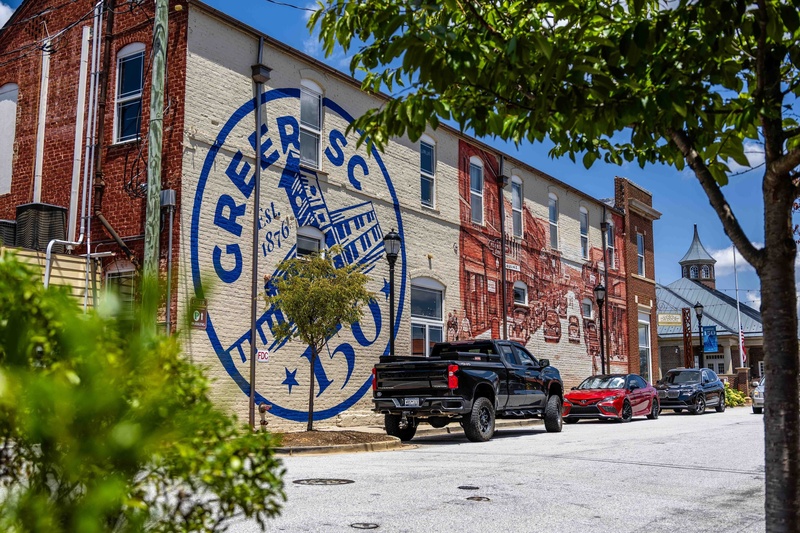 image shows a mural painted on the side of a brick building which contains the Greer150 logo and a street scene from Trade Street in 1938