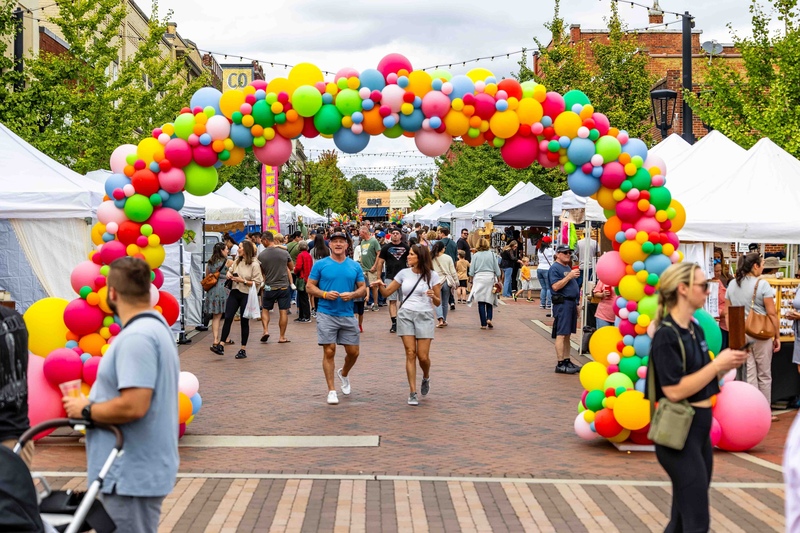 colorful balloon arch leading into the festival area for Greer Arts and Eats; you see many white event tents and dozens of people walking along brick-lined streets.
