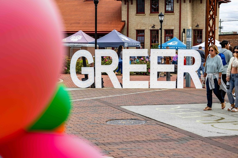 Big lighted letters spelling out GREER on the bricks of Trade Street with two ladies walking by and three balloons in the foreground