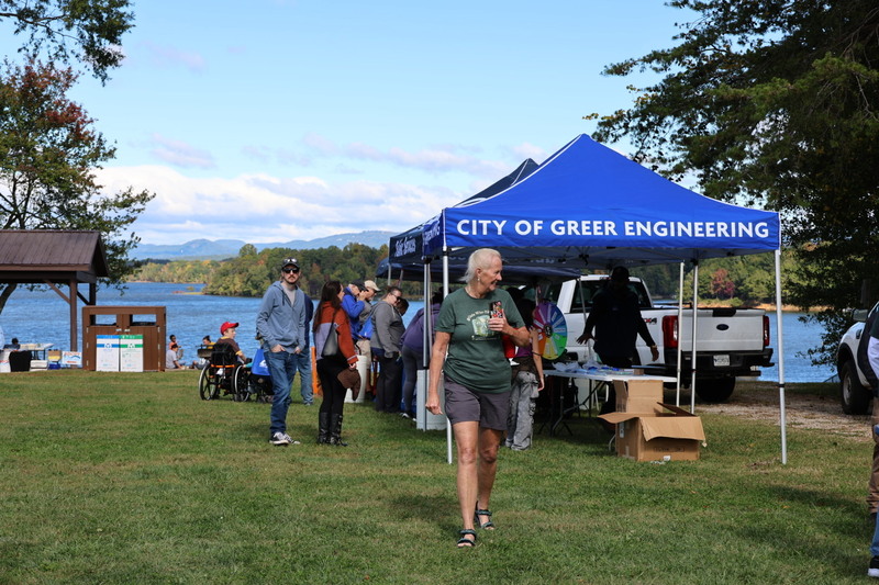 View of Lake Robinson in the background and several people gathered around blue tents on the righthand side. The one in the front says City of Greer Engineering on the canopy. A woman in shorts and a tshirt with gray hair is walking toward the camera