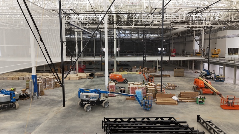 wide view of a gym under construction with pallets of wood stacked throughout and a white mesh screen hanging from the ceiling on the left side and several forklifts throughout the space