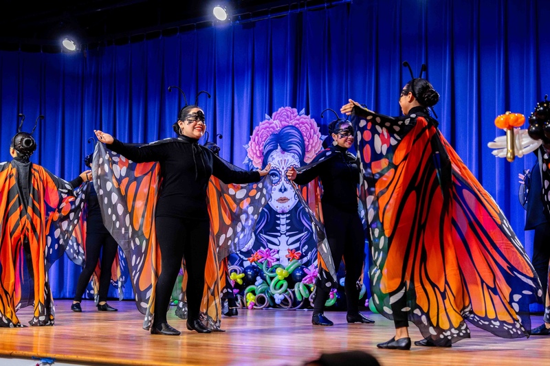 female dancers dressed as monarch butterflies dance on a stage with a day of the dead backdrop
