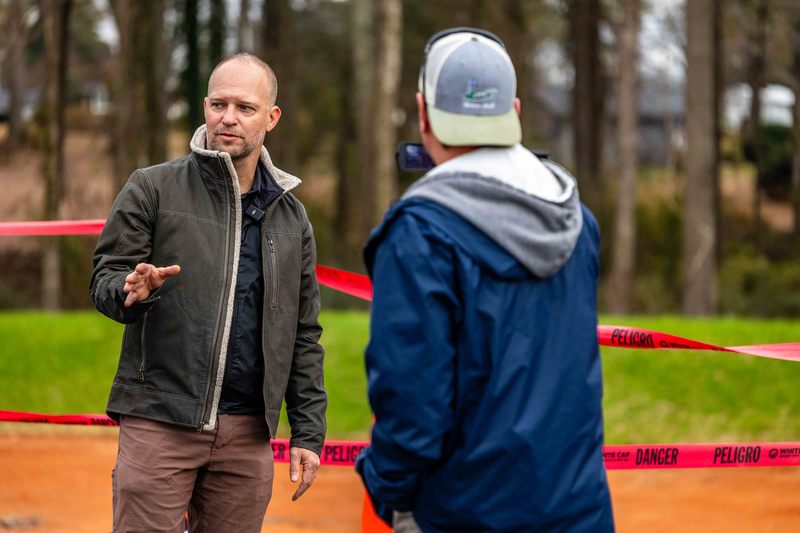 A man looks to his left with construction behind him while another man in blue records him on video