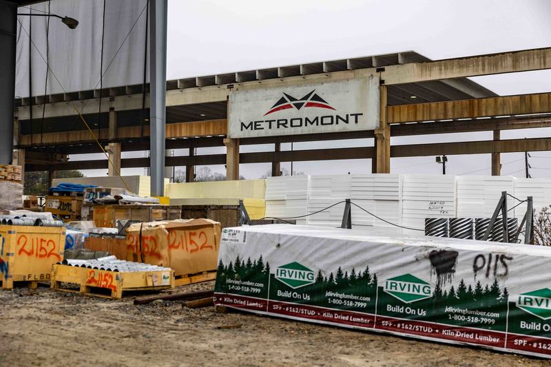 Various lumber piles and other building supplies on the ground with a concrete beam structure behind containing the Metromont logo