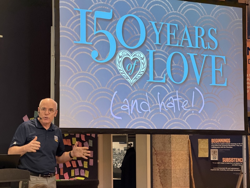 Photo shows Greer Heritage Museum director in front of a screen that says 150 years of Love and Hate