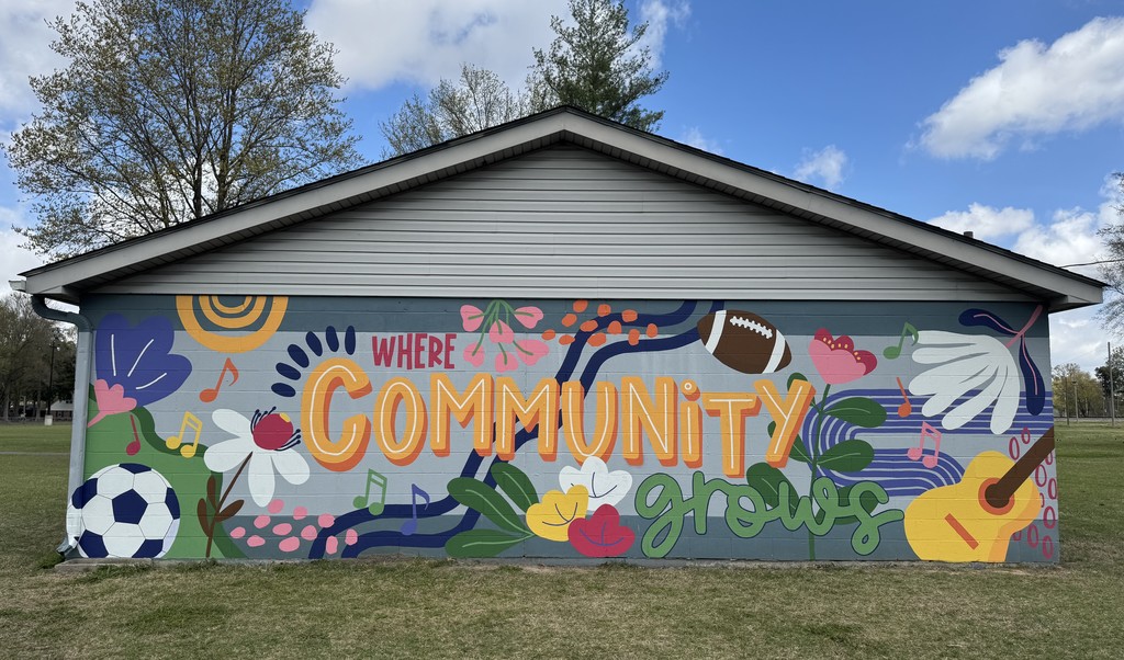 Colorful mural painted on the side of a park building at Brunetti Park. The mural reads “Where Community Grows” and features bright flowers, music notes, a football, soccer ball, and guitar against a blue-gray background under a sunny sky.