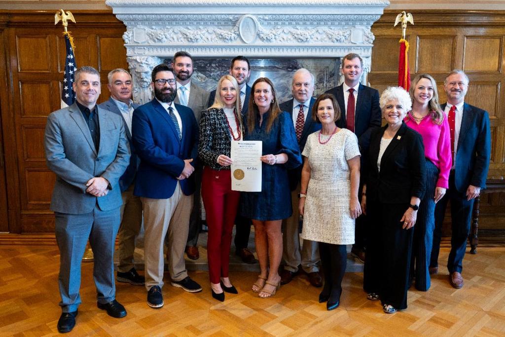 A group of community and state leaders stand together in a formal room, with two people in the center holding a signed proclamation document.