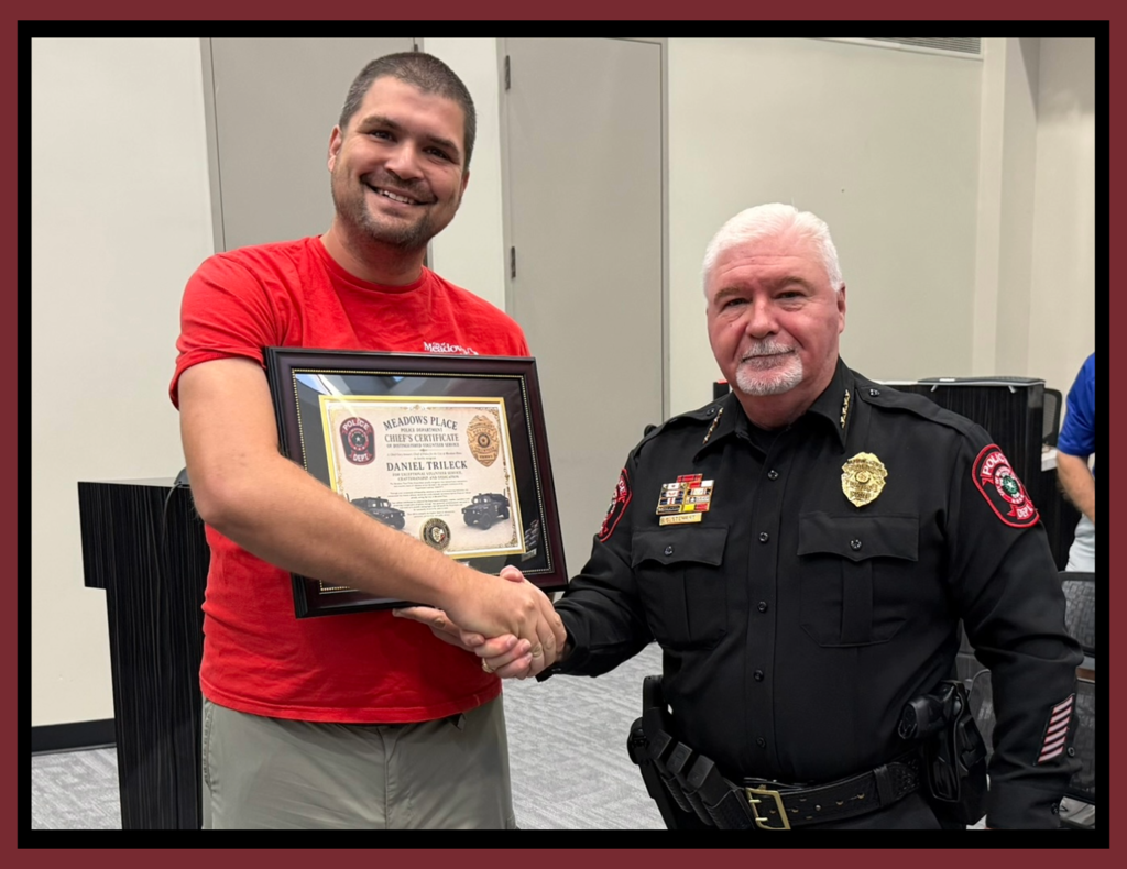 **Alt text:** Photo of Meadows Place Police Chief Gary Stewart presenting Parks and Recreation employee Daniel Trlicek with a framed Chief's Certificate of Distinguished Volunteer Service. The two shake hands during a City Council meeting while Daniel holds the certificate recognizing his contributions to the Police Department.