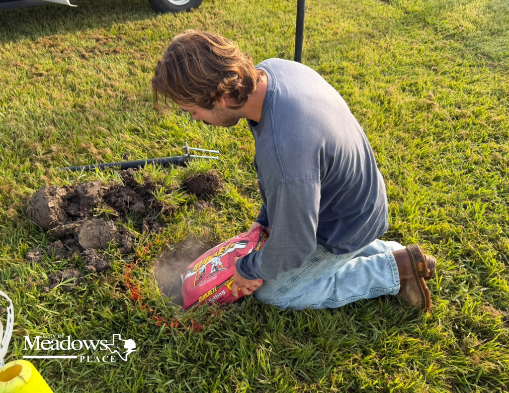 A Parks and Recreation employee kneels on the grass pouring quick-setting concrete into the base of a disc golf basket installation. Tools and freshly dug soil are visible nearby. The City of Meadows Place logo appears in the lower left corner.