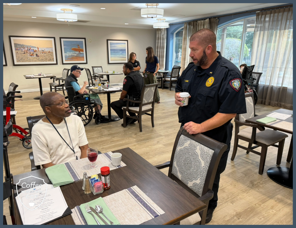 A Stafford police officer holding a coffee cup talks with a resident seated at a dining table. In the background, a Meadows Place officer visits with another resident during Coffee with a Cop.
