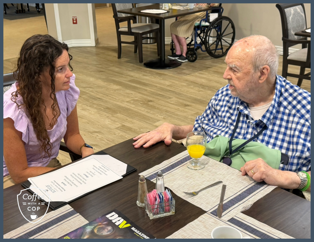 A woman in a lavender blouse talks with a resident in a blue checkered shirt seated at a dining table during Coffee with a Cop.